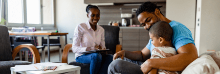A practitioner sits across from a father and his child in a living room. The practitioner smiles and takes notes.