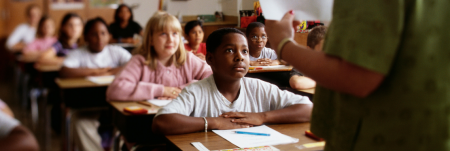 A classroom of students sit at their desk with a pencil and paper in front of them, looking up at their teacher.