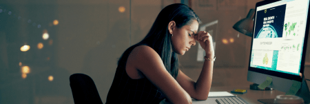 A woman sits at her computer and looks stressed. She is holding her forehead with her eyes closed.