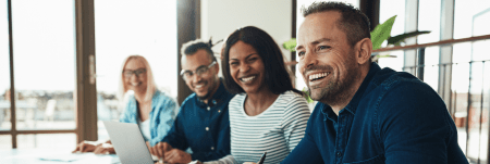 Four colleagues sit next to each other at a conference table and smile.