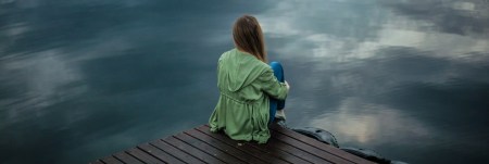 A girl sits on the edge of a dock. She faces a body of water.