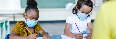 Two young girls sit at their desk in a classroom with their masks on. They concentrate on their work.