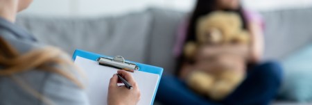 A practitioner holds up a clipboard. In the background, a young girl sits on a couch, holding a teddy bear.