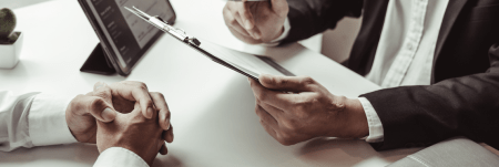 Two professionals sit at a table. One person holds up a clipboard, going over information.
