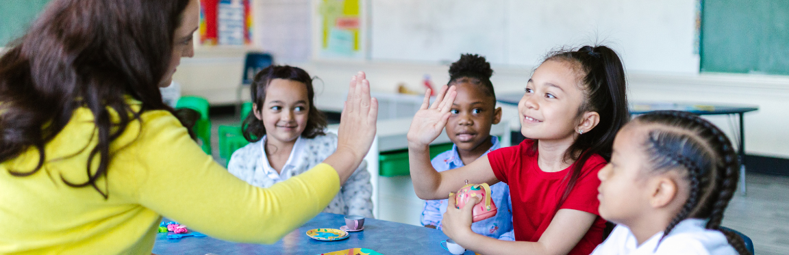 A young student sits at a table with her peers. She is high-fiving her teacher.