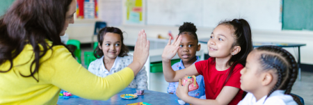 A young student sits at a table with her peers. She is high-fiving her teacher.