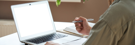 A person sits in front of a laptop screen and writes in their notebook.