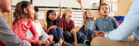 A group of children sit on the floor in their classroom next to their teacher. Two of the students raise their hand.