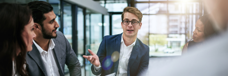 A group of professionals sit in a meeting. One person leads the conversation and the others listen.