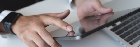 A person’s hands holding up an iPad, wearing an Apple Watch, with a laptop open in front of them.