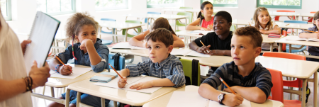 A classroom of children at their desks, listening to their teacher and writing in their notebooks.