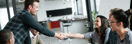 A group of people sitting down at a table in an office, while two individuals greet each other with a handshake.