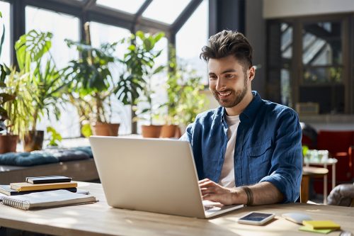 A person with short hair sits at a wooden table in a bright room filled with plants, working on a laptop and smiling. Notebooks, a smartphone, and sticky notes are scattered on the table.
