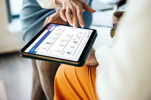 Close-up of two people interacting with a tablet device; one points at the screen displaying app icons while the other holds the tablet.